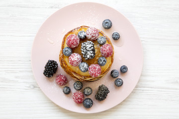 Pancakes with berries, sugar on a pink plate over white wooden background, top view. Flat lay, overhead, from above.