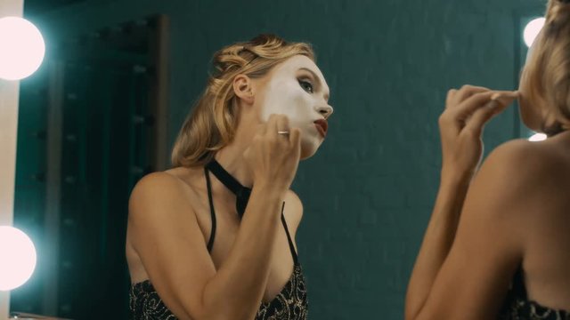 Young blond woman in dressing room doing stage makeup with white paint before performance