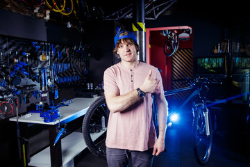 Small business and bicycle transport service. Portrait of a young man in a cap posing against the backdrop of a bicycle workshop and a tool for setting up and repairing