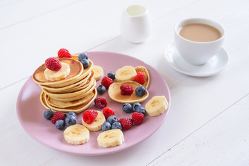 Delicious homemade pancakes with fresh berries in a violet plate on a white background. A tasty and healthy breakfast of pancakes with raspberries, blueberries and bananas. Aromatic coffee with milk.