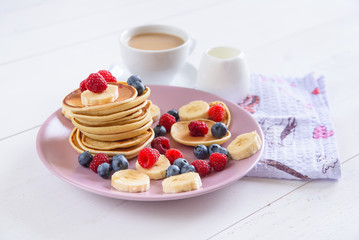 Delicious homemade pancakes with fresh berries in a violet plate on a white background. A tasty and healthy breakfast of pancakes with raspberries, blueberries and bananas. Aromatic coffee with milk.