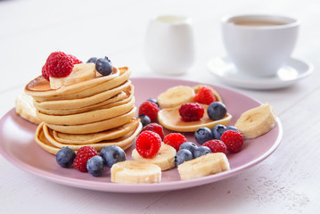 Delicious homemade pancakes with fresh berries in a violet plate on a white background. A tasty and healthy breakfast of pancakes with raspberries, blueberries and bananas. Aromatic coffee with milk.