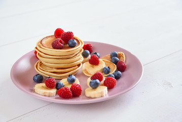 Delicious homemade pancakes with fresh berries in a violet plate on a white background. A tasty and healthy breakfast of pancakes with raspberries, blueberries and bananas.