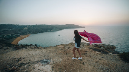 Happy young brunette woman running with pink blanket on the Calypso cave with beautiful view of Ramla bay with yellow sand beach and mediterranean sea. Gozo, Malta