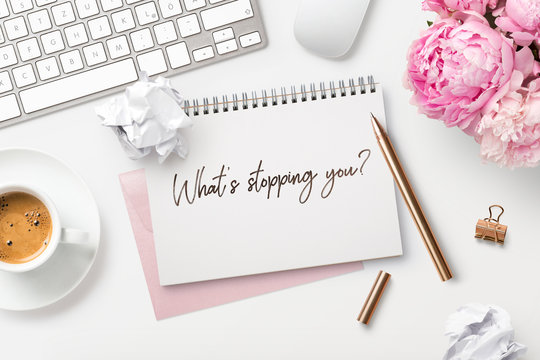 What's stopping you - business concept. Motivational quote written on a ring binder, feminine styled modern workspace with coffee, crumpled paper balls, a bunch of flowers and office gadgets, top view