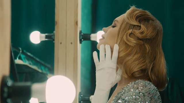 Beautiful blond woman in gorgeous sparkling dress and gloves standing in front of mirror in dressing room preparing for show