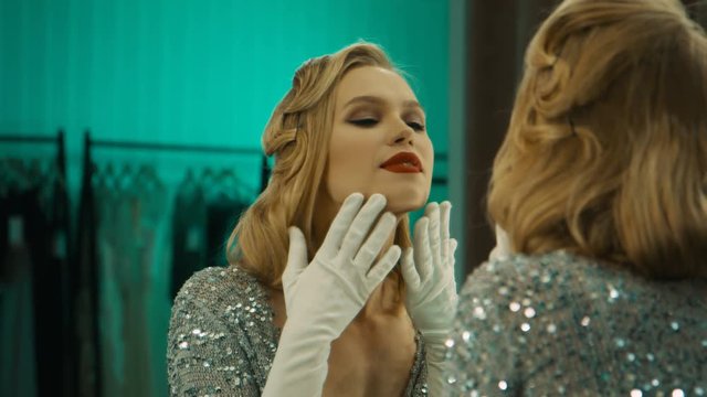 Beautiful blond woman in gorgeous sparkling dress and gloves standing in front of mirror in dressing room preparing for show
