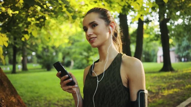 Portrait young woman use smartphone and headphone walk in park slow motion sun outdoor health hand girl tree sport fitness nature internet technology female runner exercise workout mobile cellphone