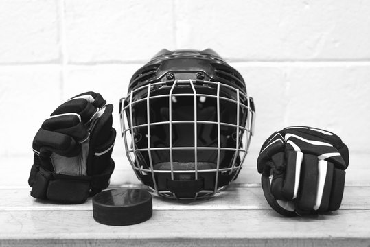 Black and white photo of  kid's hockey equipment: glows, helmet and puck. The gear are on the bench