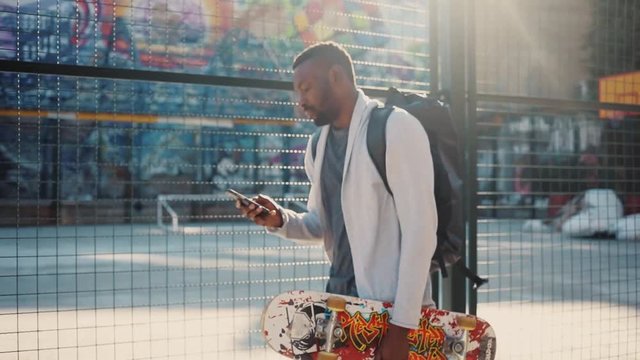 Attractive African young man in sports clothes using smartphone, chatting. Nice guy with backpack on back walking in the street. Summertime. Outdoors.