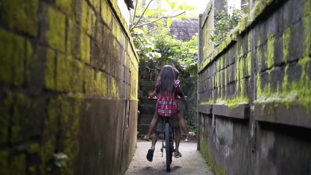 Two children bicycling during golden hour in small alley of Ubud Bali
