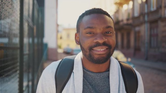 Good-looking young man with backpacks on back looking at camera, smiling. Likable African guy wearing casual clothes looking directly at camera. Outside.