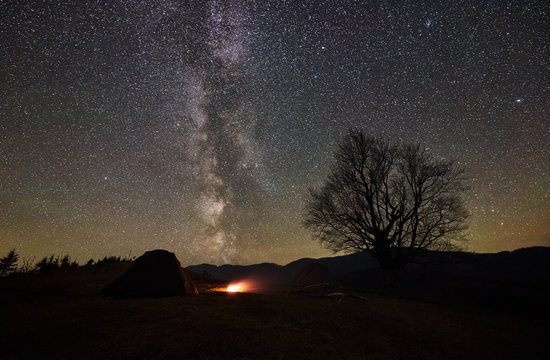 Campfire burning between two tourist tents, dark and lit from inside. Camping in mountain valley under beautiful night starry sky, Milky way. Big tree and distant mountain range on background