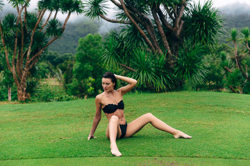 A young European-looking woman with a tanned body in a black bathing suit relaxes sitting on the green grass, straightens her hair with her hand, relaxes in the rain.