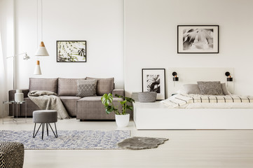 Real photo of a grey sofa standing next to a bed in monochromatic, spacious room interior with...