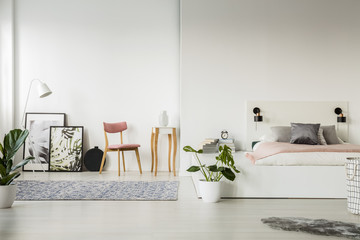 Real photo of a platform bed standing next to a chair, small table, posters and a lamp in spacious bedroom interior