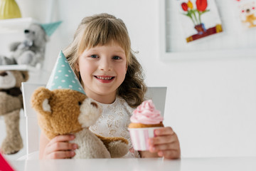 smiling birthday kid showing cupcake and holding teddy bear in cone at table