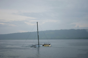 A half-sunken yacht in a misty bay. Its mast rises up against the overcast sky. A tree covered foreshore is in the distance.