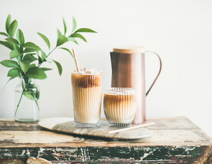 Iced coffee in glasses with milk and straws on board over rustic wooden table, white wall, jug and...