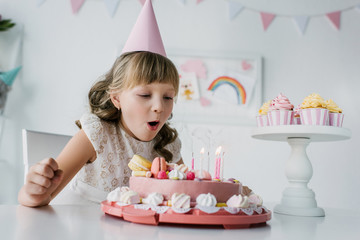 adorable kid in cone blowing out candles from birthday cake on table