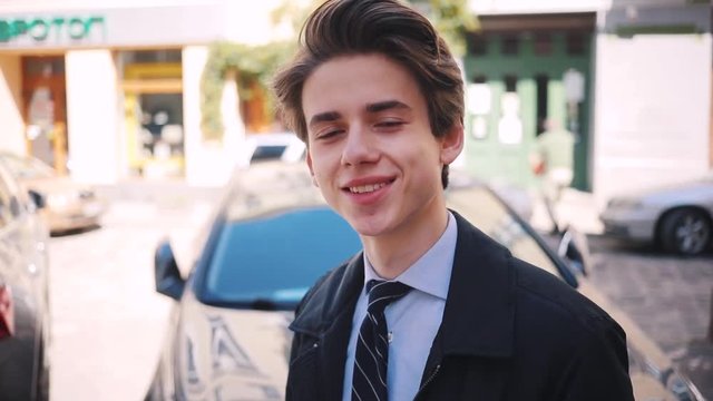 Attractive young Ukrainian man smiling and looking attentively at camera on background of cars in the street. Guy with comely haircut. Spring. Close-up.