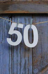 A paper cut number fifty pegged onto string across a wooden shed door painted a rustic, pastel blue , birthday card image  