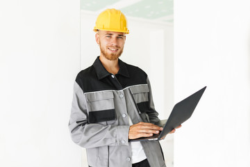 Young smiling engineer in work clothes and yellow hardhat joyfully looking in camera holding laptop in hand over white background isolated