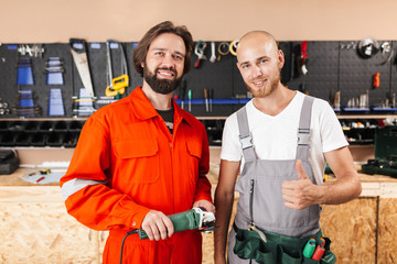 Two smiling builders in work clothes happily looking in camera spending time in workshop with tools on background