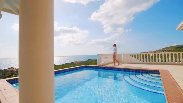 This is a very cinematic commercial use shot for travel and holidays topics. In the picture you can see female walking on a beautiful terrace near huge pool with a view to the ocean.