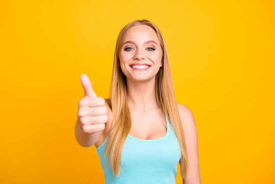 Female model with toothy beaming smile giving thumbs up success hand sign. Portrait of a charming and young blond woman isolated on yellow background