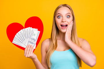 Happy beautiful girl isolated on yellow background make big toothy smile. Woman combine in one hand money and big paper carton heart figure. Concept of difficult choice