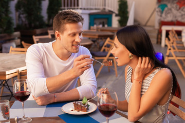 Piece of cake. Loving blonde-haired man giving some delicious piece of cake to his elegant stylish girlfriend