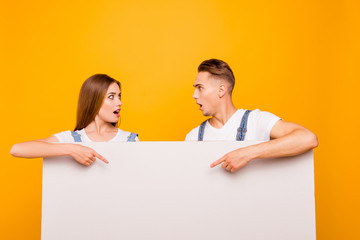 Close up portrait of funny playful lovely beautiful cute couple pointing with fingers to white blank board, showing surprising, looking astonished at each other over yellow background, isolated