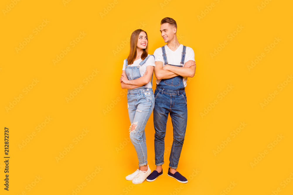 Wall mural Full lenght portrait of happy lovely adorable young cute couple with folded arms, girl standing back to guy, looking and smiling at each other over yellow background, isolated, copy space