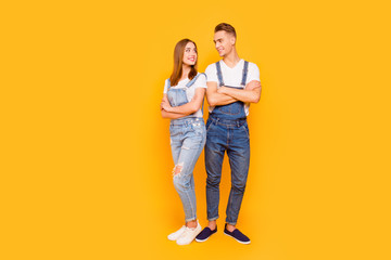 Full lenght portrait of happy lovely adorable young cute couple with folded arms, girl standing back to guy, looking and smiling at each other over yellow background, isolated, copy space
