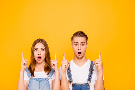 Close up portrait of lovely cheerful beautiful adorable cute lovers with wide opened mouths, pointing up with their fingers over yellow background, isolated, copy space