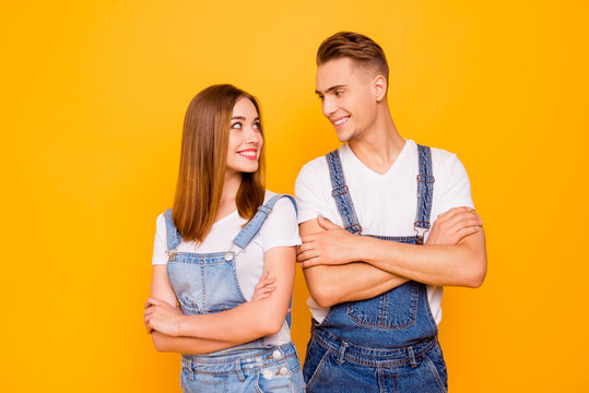 Portrait of happy lovely adorable young cute couple with crossed arms, girl standing back to guy, looking and smiling at each other over yellow background, isolated