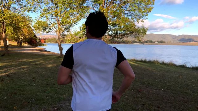 We follow a runner as he jogs along a lake. Setting beautiful lake side with natural trees and mountain surroundings.