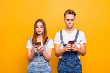 Portrait of cute young couple wearing casual, holding their smartphones in hands, seriously looking at screens, over yellow background, isolated