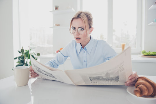 Adult adorable attractive astonished woman office executive worker wearing eyeglasses keeping in hands and reading newspaper early in the morning having a drink with croissant in light kitchen