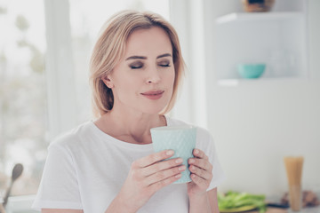 Close up portrait of adorable cheerful smiling young woman with nice manicure  keeping glass cup of drink in hands with closed eyes wearing white t-shirt