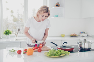 Charming adorable attractive woman wearing pajama chopping with knife red pepper on board in kithcen. Tomatoes, salad, cucumber, pan, pot, yellow pepper lying on table