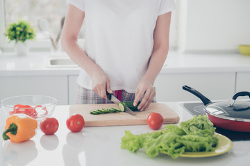Attractive beautiful charming female manicure hands chopping green fresh cucumber  on board with knife, cooking nice salad. Chopped pepper, tomatoes, yellow pepper lying on white table in kitchen