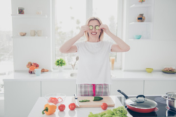 Attractive adorable beautiful charming nice smiling blonde woman wearing pajama putting chopped pieces of cucumber to her eyes having fun while cooking salad