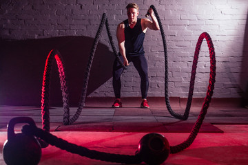 young handsome white man in black t-shirt is engaged with ropes in the gym on a black brick background. The idea of cardio training