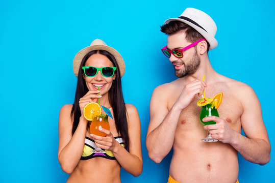 Beach style! Closeup portrait of handsome young man with beard holds drink in his hand and looks on charming girl which is drinking cocktail isolated on blue background