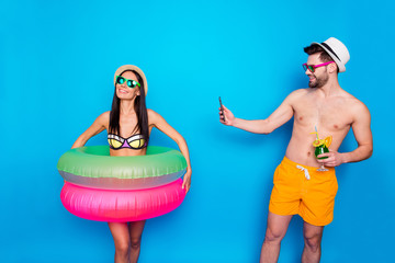 Guy in the hat, sunglasses and bright shorts holding a cool cocktail and taking pictures of a happy girl with multi-colored inflatable circles on the smartphone