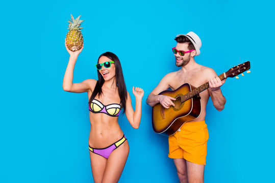 Time to celebrate vacations! Brunette guy with beard plays on guitar in hot summer day while a beautiful and charming girl dances to his music with pineapple in her hand isolated on blue background