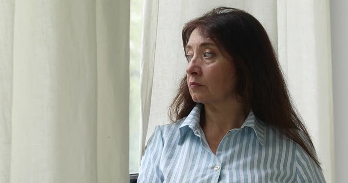 Serious adult pensive woman in striped shirt and with long brown hair standing near window with curtain looking away