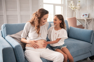 Touching belly. Excited girl wearing white open shoulder blouse touching belly of her pregnant aunt